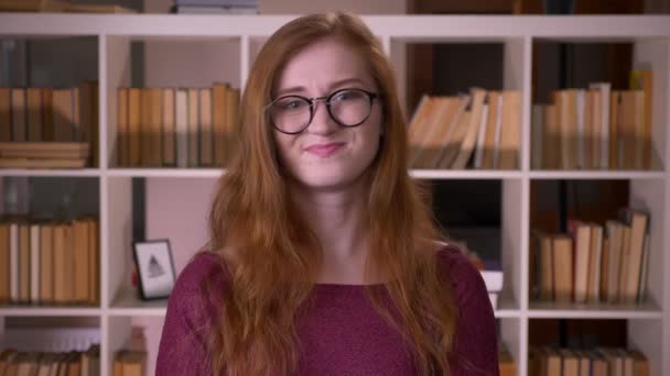大学の図書館でカメラを見ないで頭を振るメガネをかけた若い赤毛の魅力的な白人女子学生のクローズアップ肖像画 — ストック動画