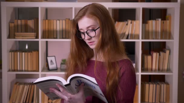 Крупный план портрета молодой рыжей привлекательной кавказской студентки в очках, читающей книгу, смотрящей в камеру и улыбающейся в библиотеке колледжа — стоковое видео