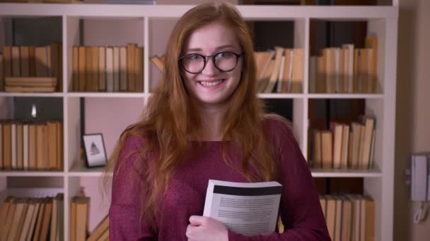 Close-up retrato de jovem ruiva atraente caucasiana estudante em óculos segurando um livro olhando para a câmera e sorrindo na biblioteca da faculdade — Vídeo de Stock