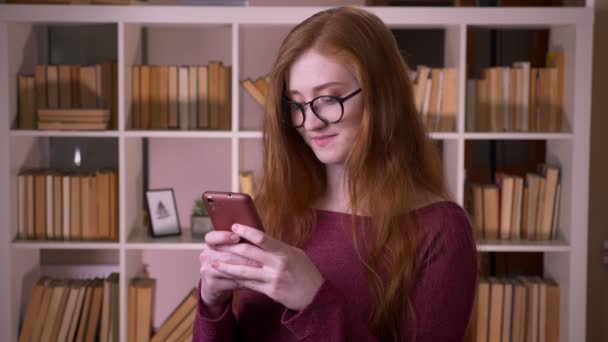 Close-up retrato de jovem ruiva atraente caucasiana estudante em óculos usando o telefone sorrindo alegremente na biblioteca da faculdade — Vídeo de Stock
