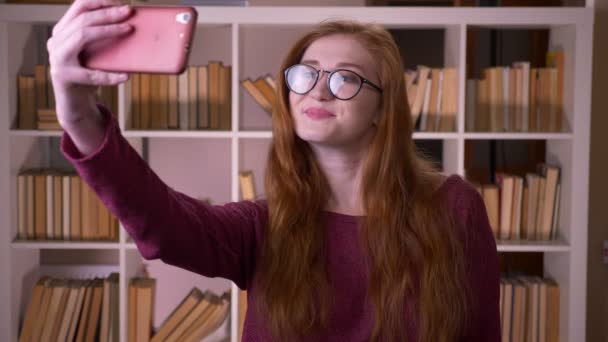 Крупный план портрета молодой рыжей привлекательной кавказской студентки в очках, делающей селфи по телефону в библиотеке колледжа — стоковое видео