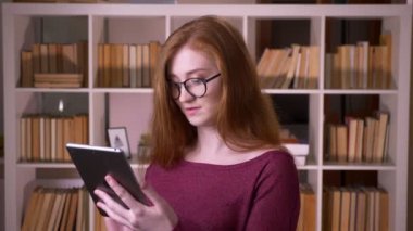 Üniversite kütüphanesinde tablette gözlükle gezinen genç kızıl saçlı çekici beyaz kız öğrencinin yakın çekim portresi