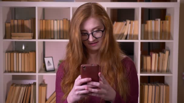 Closeup portrait of young redhead attractive caucasian female student in glasses browsing on the phone in the college library — Stock Video