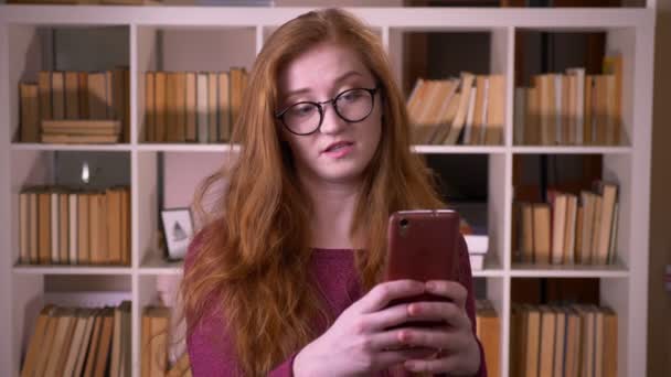 Closeup portrait of young redhead attractive caucasian female student in glasses having a video call on the phone in the college library — Stock Video