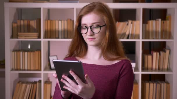 Primer plano retrato de la joven pelirroja atractiva estudiante caucásica en gafas usando la tableta y mostrando la pantalla verde a la cámara en la biblioteca de la universidad — Vídeos de Stock