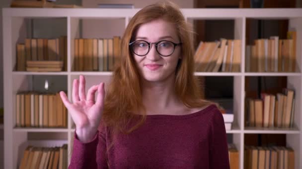 大学図書館のカメラを見て大丈夫な看板を示す眼鏡で若い赤毛魅力的な白人女子学生のクローズアップ肖像画 — ストック動画
