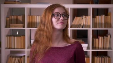 Üniversite kütüphanesinde kamera önünde neşeli dans gözlüklü genç kızıl saçlı çekici beyaz kız öğrencinin closeup portresi