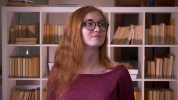 Närbild porträtt av unga rödhårig attraktiv kaukasiska kvinnlig student i glasögon dansa glatt framför kameran i kollegiet biblioteket — Stockvideo