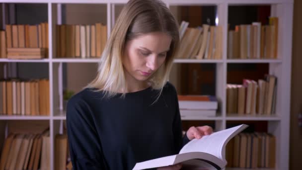 Sesión de primer plano de una joven estudiante atractiva leyendo un libro mirando a la cámara en el interior de la biblioteca universitaria — Vídeo de stock