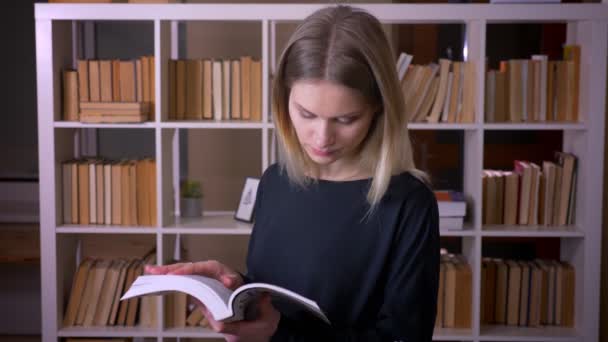Съемки крупным планом молодой привлекательной студентки, читающей книгу, смотрящей на камеру и счастливо улыбающейся в помещении университетской библиотеки — стоковое видео