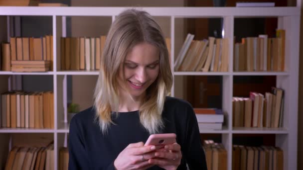 Sesión de primer plano de la joven estudiante atractiva utilizando el teléfono mirando a la cámara sonriendo felizmente en el interior de la biblioteca de la universidad — Vídeo de stock