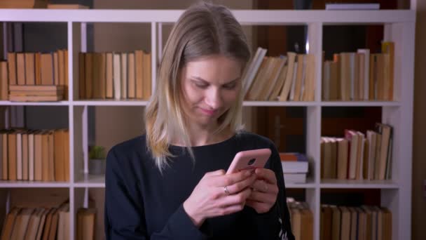 Sesión de cerca de jóvenes atractiva estudiante mensajes de texto en el teléfono mirando a la cámara sonriendo alegremente en el interior de la biblioteca de la universidad — Vídeo de stock