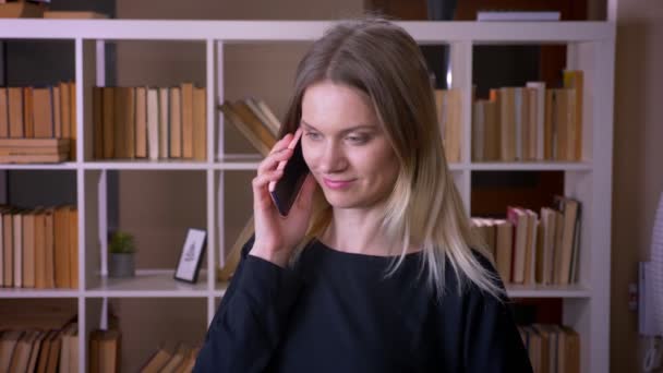 Sesión de primer plano de una joven estudiante atractiva que tiene una llamada telefónica en el interior de la biblioteca universitaria — Vídeo de stock