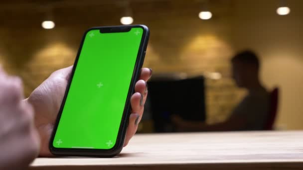 Gros plan vue arrière de la main féminine jouant une vidéo sur le téléphone avec écran chromatique vert le tenant verticalement dans le bureau à l'intérieur — Video