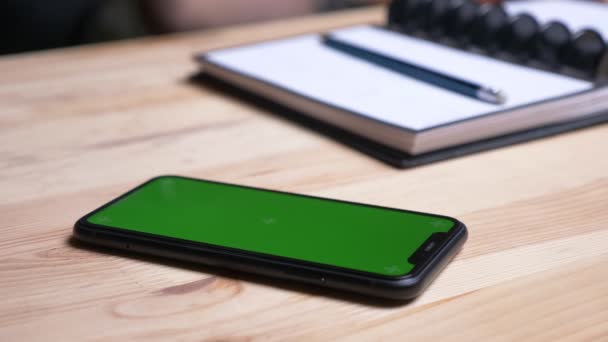 オフィス内のノートの横の机の上に置く緑色の画面で携帯電話を取る女性の手のクローズアップ撮影 — ストック動画