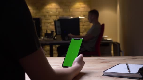 Vue arrière rapprochée prise de vue de la main féminine tenant un téléphone avec écran chromatique vert dans le bureau à l'intérieur avec des employés sur le fond 