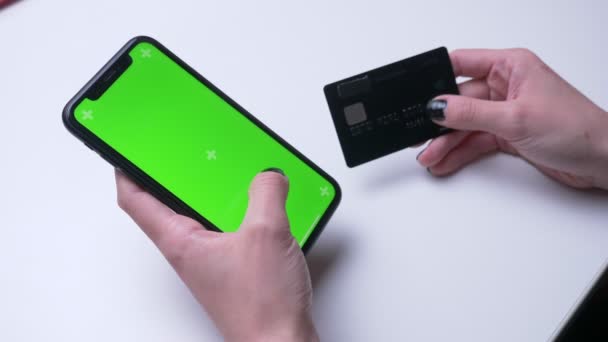 Closeup back view shoot of female hand shopping online on the phone with green chroma screen holding a credit card in the office indoors on the white desk — Stock Video