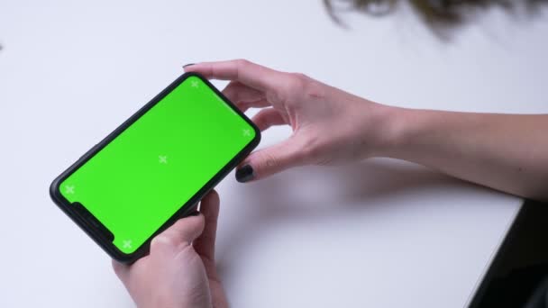 白い机の上のオフィスの屋内で緑色のクロマスクリーンで携帯電話でビデオ広告を再生する女性の手のクローズアップバックビュー撮影 — ストック動画