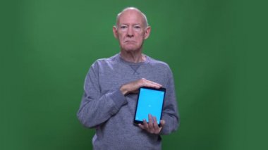 Tableti kullanan ve yeşil üzerinde izole arka plan ile kamera bakarak mavi ekran gösteren eski üst düzey kafkas adam Closeup ateş