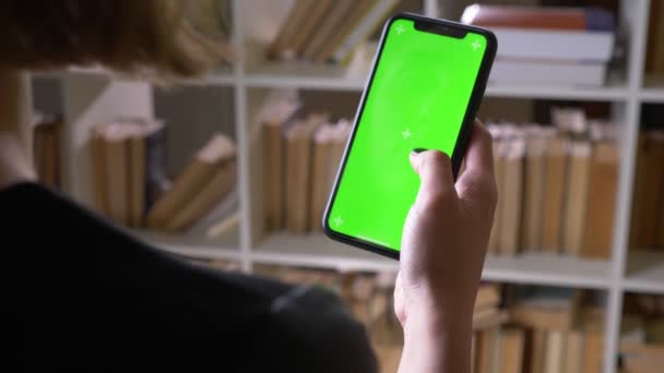 Крупный план Задний вид съемки студенток вручную прокручивания по телефону с зеленым хроматическим экраном в библиотеке в помещении — стоковое видео