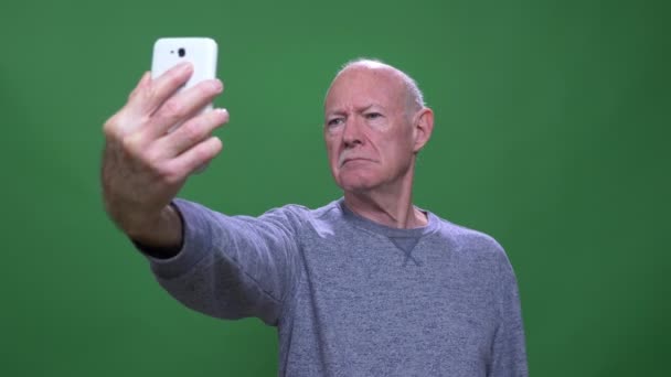 Disparo de primer plano del viejo hombre caucásico mayor tomando selfies en el teléfono mirando a la cámara con fondo aislado en verde — Vídeo de stock