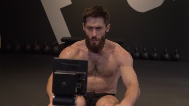Lukke direkte opptak av voksen, atletisk skjorteløs mann med romaskin og gjøre motstandstrening svettende innendørs i gymsalen – stockvideo