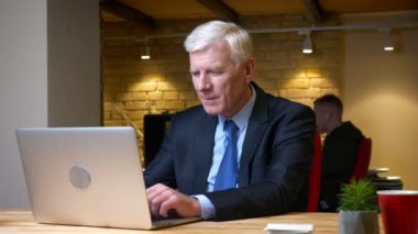 Dizüstü bilgisayarı kullanan ve işyerinde ofiste ki kameraya bakarak gülümseyen eski beyaz işadamının yakın çekim çekimi