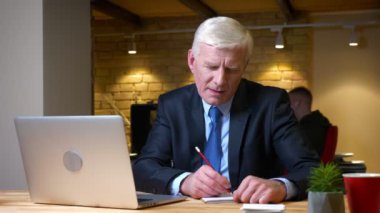 Dizüstü bilgisayarı kullanarak eski beyaz işadamının yakın çekim çekimi not alıyor ve işyerinde ofiste ki kameraya mutlu bir şekilde bakıyor