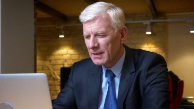 Eski başarılı kafkas işadamının dizüstü bilgisayarı kullanarak ve iş yerinde ofiste mutlu bir şekilde gülümseyerek kameraya parmağını göstererek yakın çekim