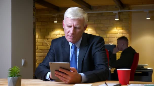 Closeup shoot of old caucasian businessman browsing on the tablet and drinking coffee indoors in the office on the workplace — Stock Video