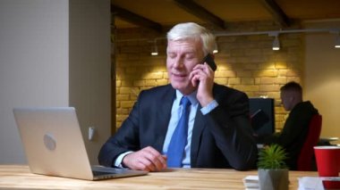 İşyerinde ofiste ki dizüstü bilgisayarı kullanırken telefonda gülümseyerek sohbet eden eski beyaz işadamının yakın çekim çekimi