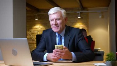 İşyerinde ki ofiste ki dizüstü bilgisayarın önünde mutlu bir şekilde gülümseyerek para gösteren eski beyaz işadamının yakın çekim çekimi