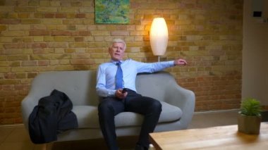 Eski beyaz işadamının ceketini çıkarıp dairenin içinde kanepede oturan televizyonu seyreden yakın çekim portresi