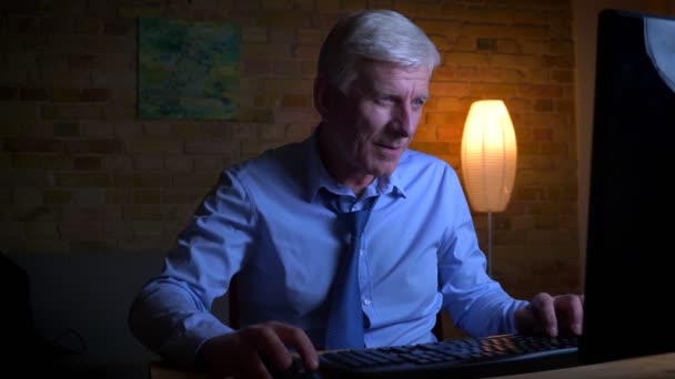 Портрет крупным планом старого кавказского бизнесмена, играющего в видеоигры на компьютере в помещении квартиры с выключенным светом — стоковое видео