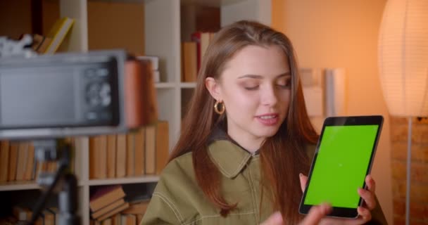 Female vlogger shows green chroma screen of tabet into camera and talking about it on bookshelves background. — Stock Video