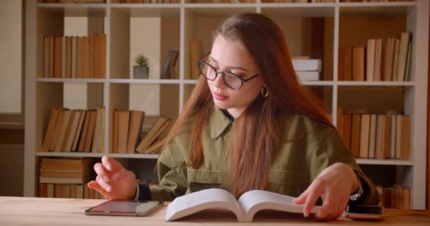 Ritratto di giovane studentessa che legge un libro e cerca informazioni in tablet facendo i compiti in biblioteca . — Video Stock