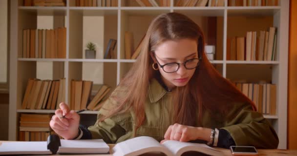 Retrato de una joven estudiante leyendo un libro y escribiendo tareas en relojes de copybook en la cámara de la biblioteca . — Vídeo de stock