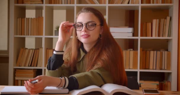 Retrato de una joven estudiante sentada con relojes de libros en la cámara con atención en la biblioteca . — Vídeo de stock