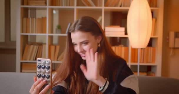 Junge Teenager-Bloggerin spricht im Videochat auf dem Handy auf dem Sofa auf Bücherregalen Hintergrund zu Hause sitzen. — Stockvideo