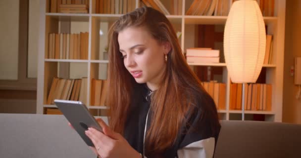 Porträt eines jungen Teenager-Mädchens, das mit Tablet arbeitet, sitzt auf dem Sofa im Bücherregal und schaut zu Hause in die Kamera. — Stockvideo