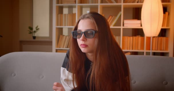 Portrait of young teenage girl in 3d glasses watches film on TV sitting on sofa on bookshelves background at home. — Stock Video