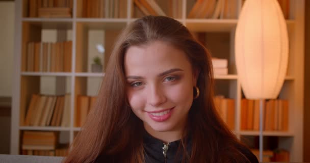 Close-up portrait of young female student smiles into camera seductively on bookshelves background. — Stock Video