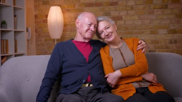 Esposos caucasianos sênior sentados juntos no sofá abraçando uns aos outros sorrindo lindamente para a câmera em casa . — Vídeo de Stock