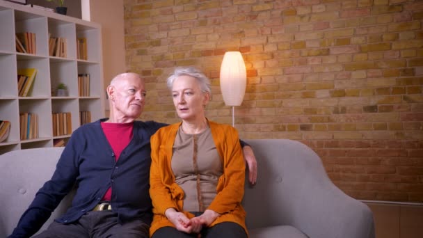 Senior kaukasiska makar sitter tillsammans vid soffan krama och prata med andra vara lycklig hemma. — Stockvideo