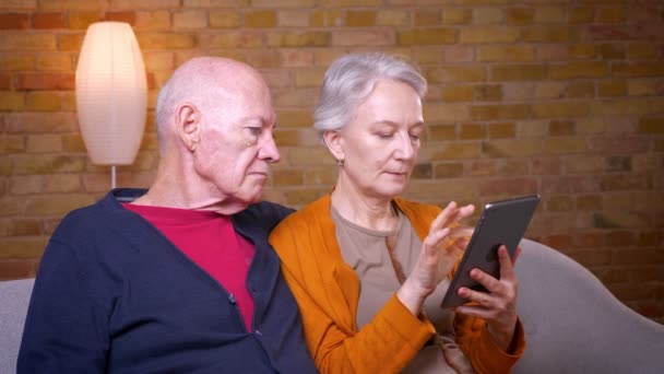 Ältere kaukasische Eheleute schauen aufmerksam ins Wohnzimmer, wo sie auf dem Sofa sitzen und einander umarmen. — Stockvideo