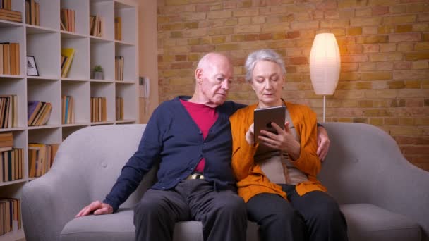 Les conjoints caucasiens aînés regardent en tablette être joyeux et heureux assis sur le canapé dans le salon . — Video