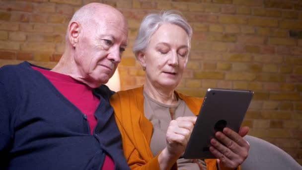 Esposos caucasianos seniores usando o aplicativo juntos no tablet sentado no sofá na sala de estar . — Vídeo de Stock