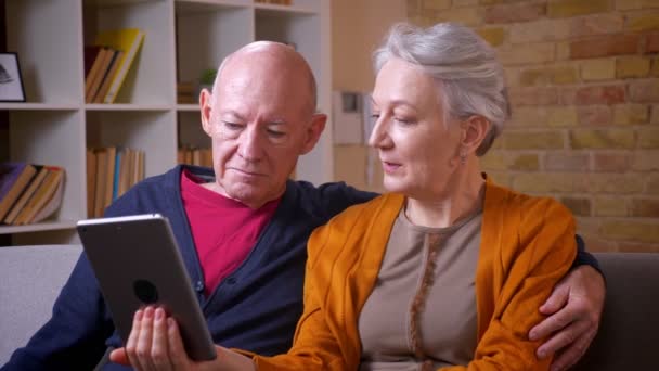 Äldre kaukasiska makar tittar på tabletten och disussing det är positivt och glad på soffan i vardagsrummet. — Stockvideo