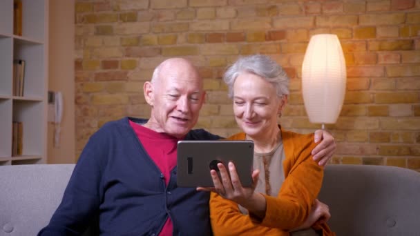 Two senior gray-haired caucasian spouses having a videocall on tablet being funny and joyful at cozy home. — Stock Video