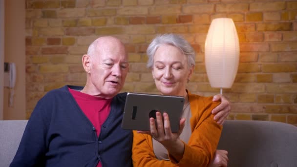 Senior gray-haired caucasian couple having online videocall on tablet being happy and joyful at cozy home. — Stock Video